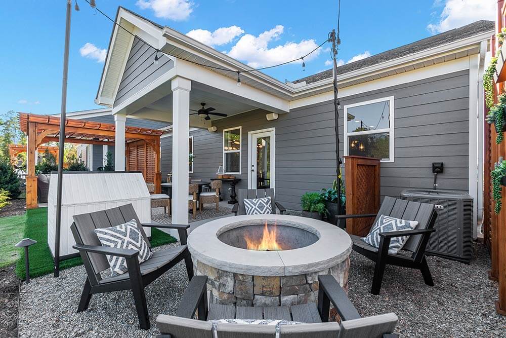 A cozy fall backyard featuring a circular fire pit surrounded by modern patio chairs with patterned cushions, warm string lights overhead, and a wooden pergola, creating the perfect space for autumn gatherings and s'mores under the crisp fall night sky. 