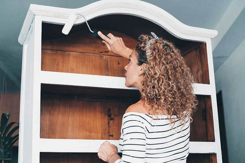 A woman transforms a wooden bookshelf with a coat of white paint, capturing the spirit of spring DIY home improvement. This project exemplifies how a little creativity and effort can bring new life to existing furniture without overspending.