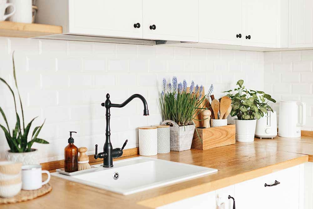 A beautifully styled kitchen countertop with potted herbs, Muscari flowers, and wood-toned accessories brings spring's essence into the heart of the home. The clean white cabinets, ceramic sink, and natural textures reflect 2025 decor trends focusing on fresh, nature-inspired spaces.